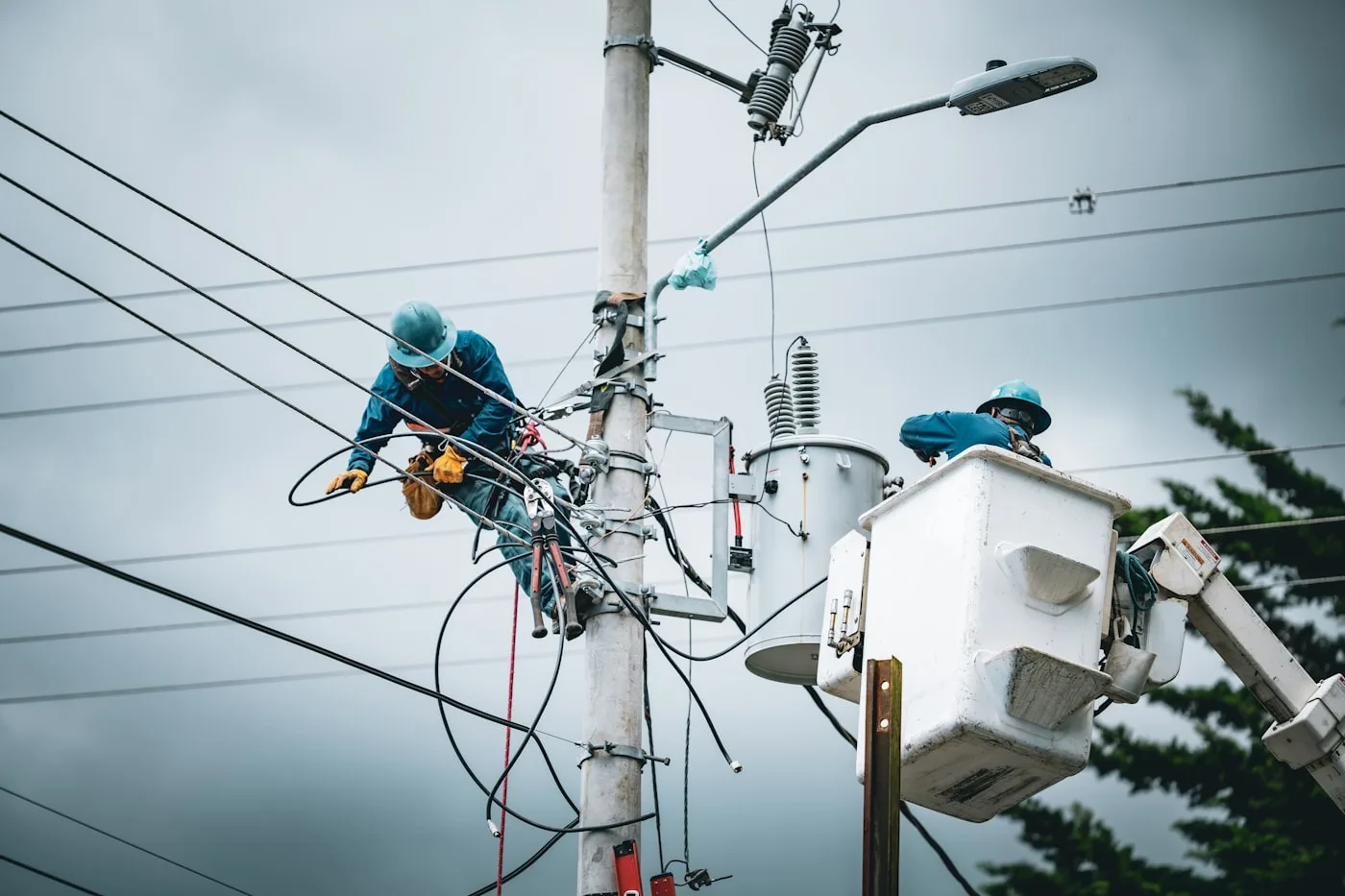 Two utility workers in a bucket truck performing maintenance on a medium-voltage distribution pole under an overcast sky